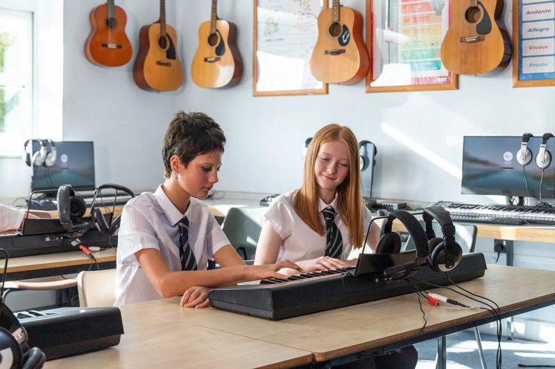 Two girls are sitting side by side playing an electronic keyboard together in a classroom music lesson. The scene captures collaboration and shared learning in a creative and supportive educational environment.