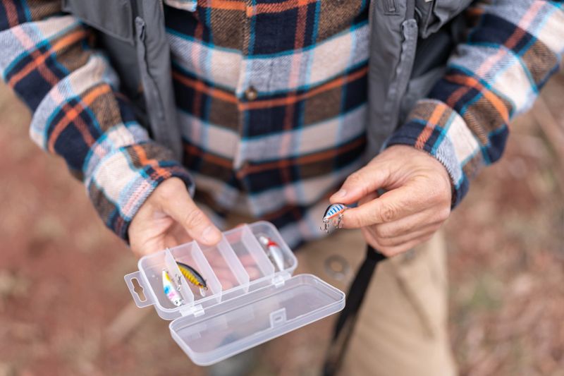 A close-up of the hands of a senior fisherman stands outdoors in nature, selecting lures from a clear tackle box, holding a rod. He is dressed in a plaid shirt with a fishing vest over it.