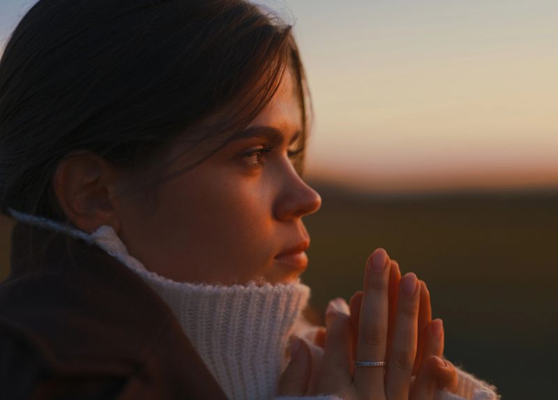 Close-up profile portrait of young woman with hands folded during warm sunset light. She wears white sweater and expresses thoughtful, hopeful, and serene mood outdoors.