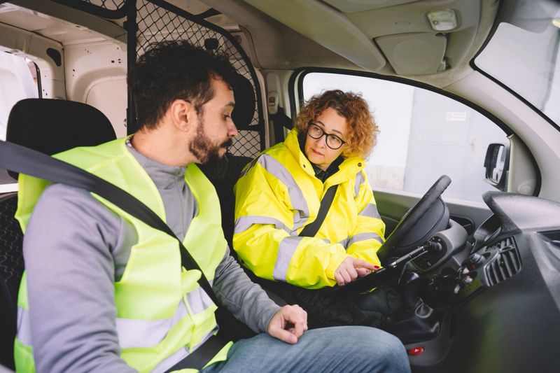 Two colleagues are engaged in a discussion inside a delivery van, focused on planning their logistics route. This interaction emphasizes collaboration and communication, essential elements for enhancing workflow in the delivery and transport industry.