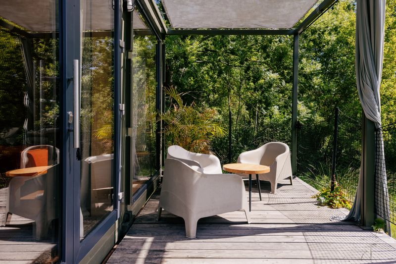 Modern house with garden. Outdoor seating area features two white chairs and a small wooden table. Terrace or patio space is bright and inviting, surrounded by lush greenery and natural light.
