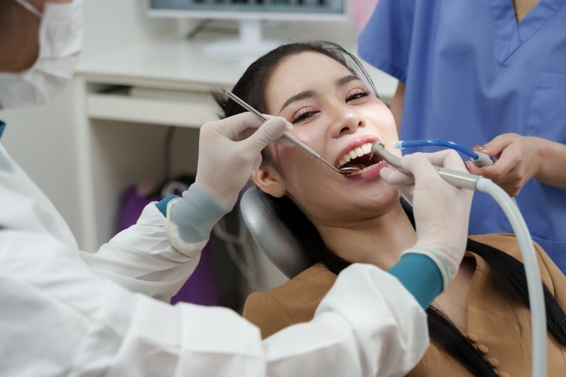 Asian adult female patient smiling while receiving oral cleaning treatment in dental clinic, dentist uses scaler and suction equipment, dental assistant present in background during procedure