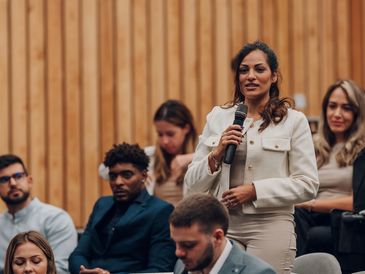 A woman speaks confidently with a microphone in a professional audience setting.