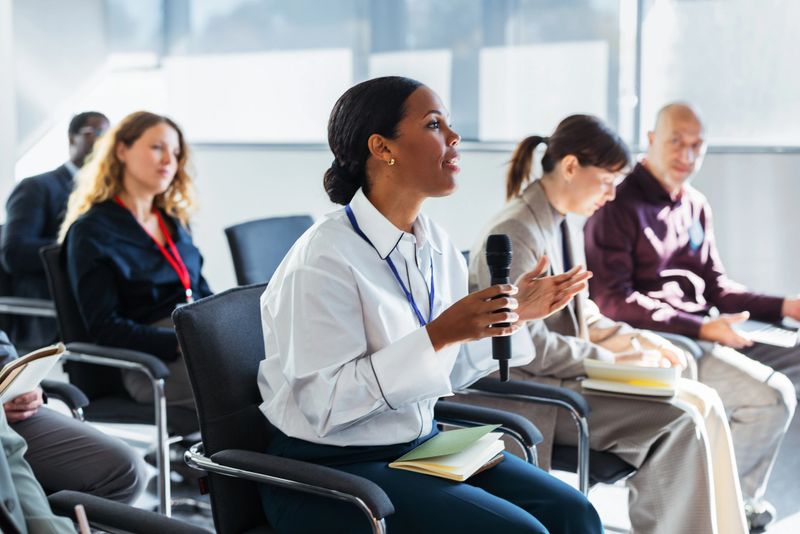 A confident professional woman speaks into a microphone while peers listen, take notes, and engage in a collaborative discussion in a bright, modern conference room.