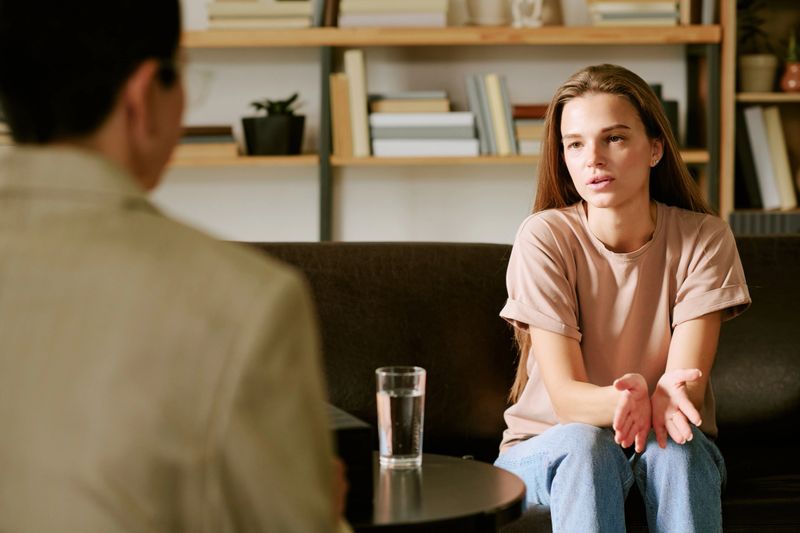 Young adult Caucasian woman sitting on sofa talking to middle aged Caucasian man during psychological counseling session, gesturing with hands, glass of water on table