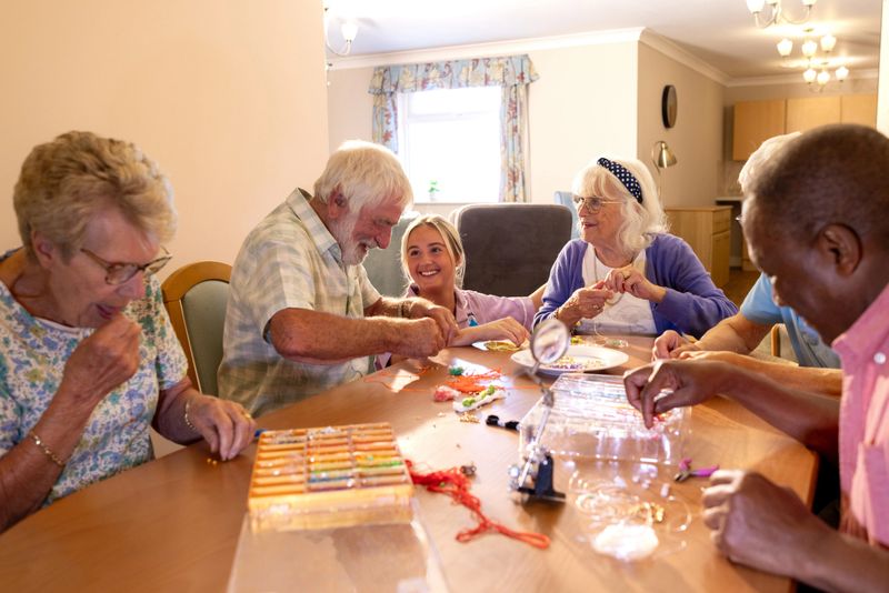 A wide shot of a group of elderly adults, sitting together, participating in a crafting session around a wooden table in a well-lit, cosy indoor setting. A young professional healthcare worker checks on them, smiling. Located in Durham, England.