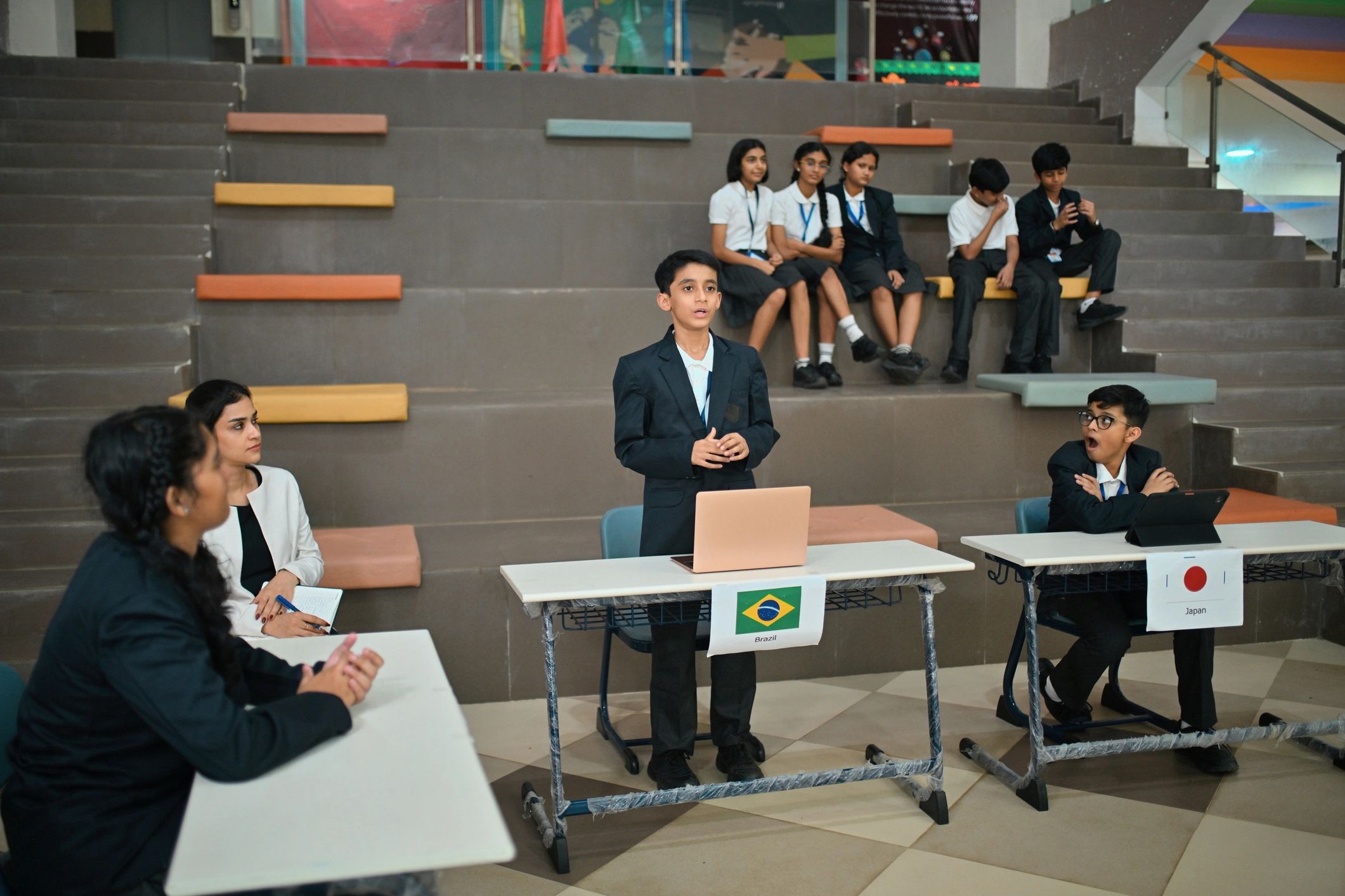Middle school students act as country delegates for Brazil, Japan, and the United Arab Emirates, presenting and discussing at their laptops while their classmates watch from the tiered seating behind them, recreating the atmosphere of a real global summit.