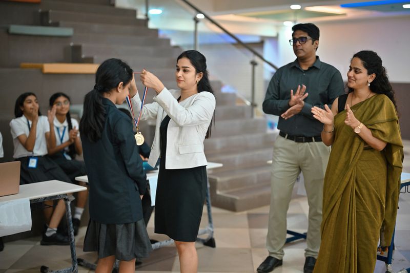 A student stands proudly as a school administrator places a gold medal around her neck during an awards ceremony. Teachers and classmates stand nearby, clapping and celebrating her achievement. The moment captures pride, recognition, and encouragement as the school community applauds her success.