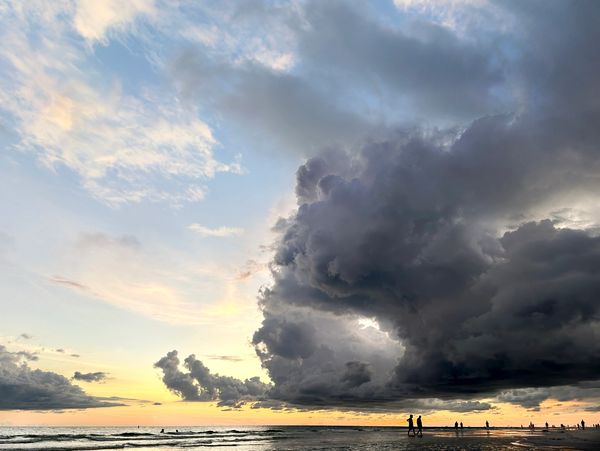 Dramatic dark clouds gather over a sunset beach with silhouettes of people walking.