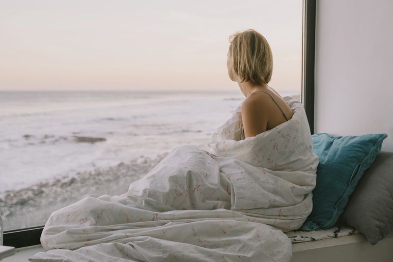 Thoughtful woman relaxes by the window. Woman sitting by panoramic window with ocean view, wrapped in blanket enjoying morning. Serene female in blanket sits on the windowsill with sea view.