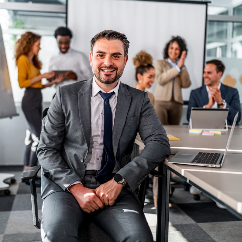 Confident male executive smiling at camera while diverse business team claps, celebrating success at an office meeting