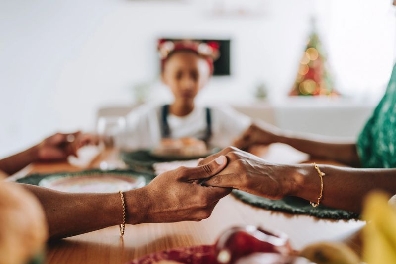 Close-up of family members holding hands at the dining table before a Christmas meal. Soft focus on a child in the background and a decorated Christmas tree. Concept of unity, gratitude, holiday tradition, emotional connection and family support