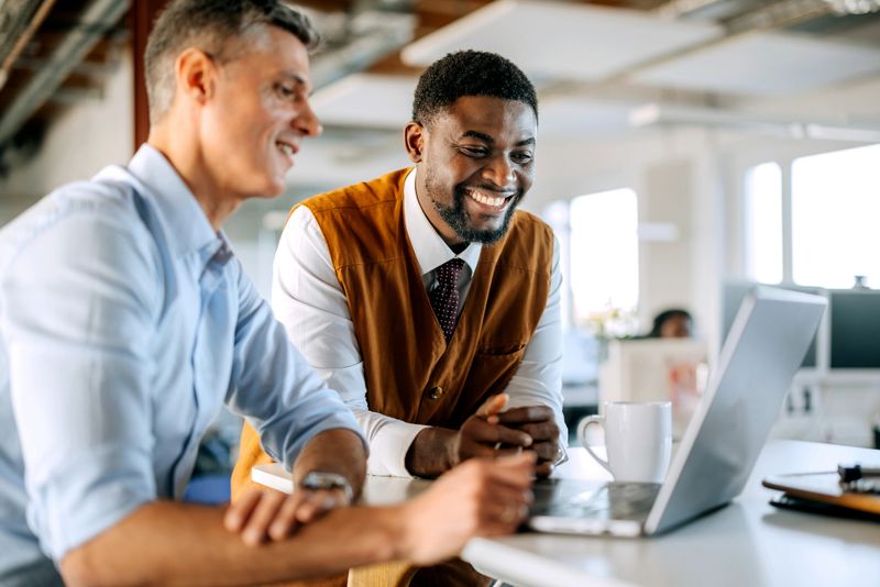 Two male colleagues of diverse backgrounds collaborate in an office, discussing work while using a laptop. Captures teamwork, professional communication, and a modern workplace environment