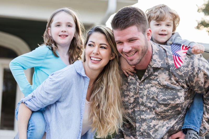 Soldier embraces family after return. Daughter and son hold flag, sharing happiness and togetherness.