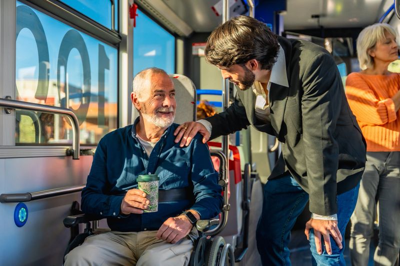 Young man assisting a smiling senior man in a wheelchair boarding a city bus, offering supportive, kind help during a sunny urban commute with warm natural light