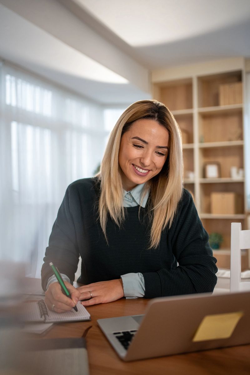 Smiling young professional woman focused on writing in a notebook while working remotely from her cozy home office with a laptop on a wooden desk, planning and studying confidently