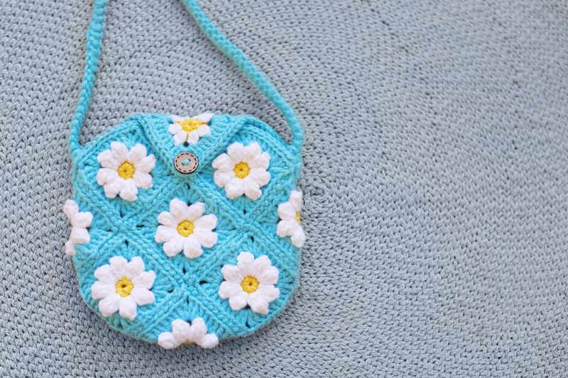 Overhead close-up of small, round, handmade blue crocheted shoulder bag featuring daisy flower pattern on turquoise background, resting on knit surface