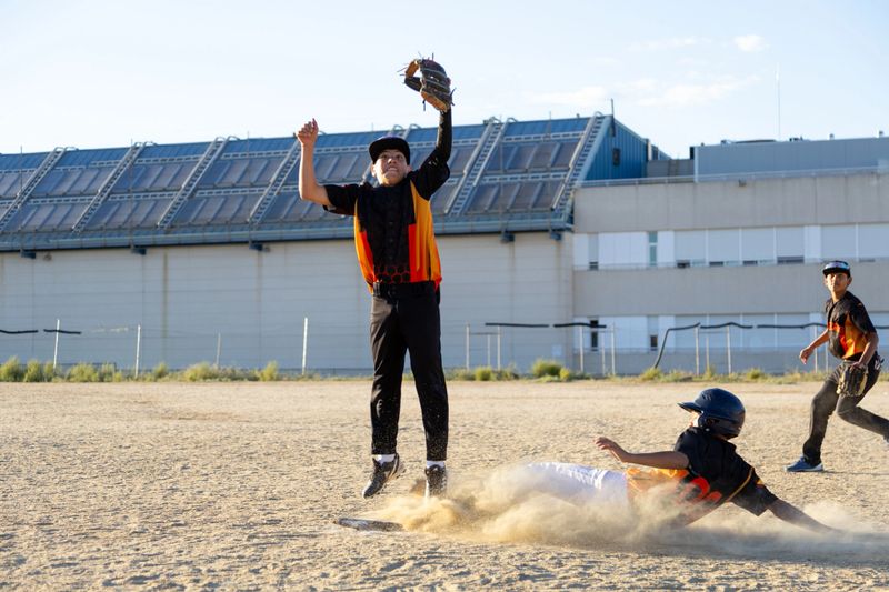 Kids sliding into base creating dust while another player jumps to catch the ball during a youth baseball game