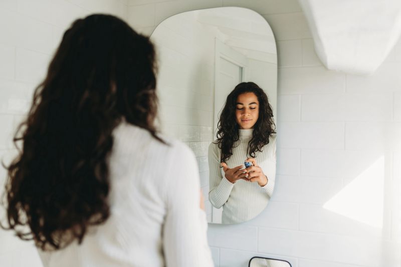 Woman standing in bathroom applying a skincare serum from a dropper bottle to her hand, practicing a morning self-care routine