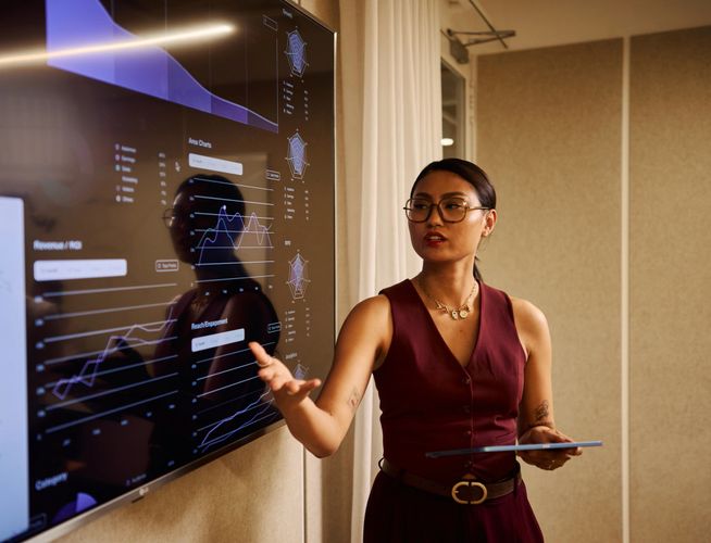 A woman presents data charts on a large screen during a business meeting.