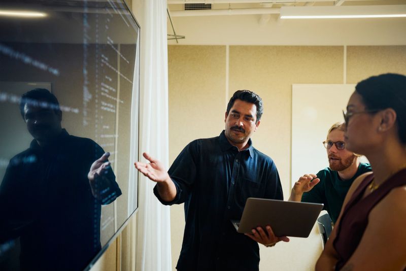 Woman pointing at code on a large screen while discussing with a colleague in an office