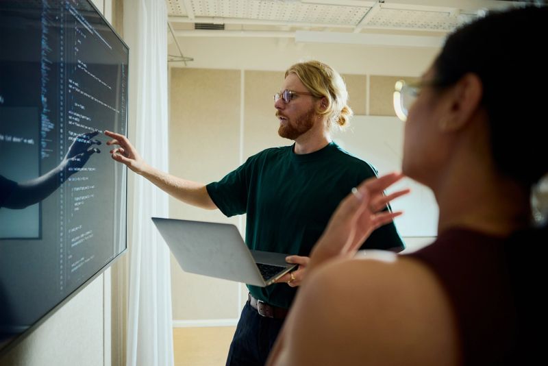 Programmer holding laptop, showing software code on large screen in an office