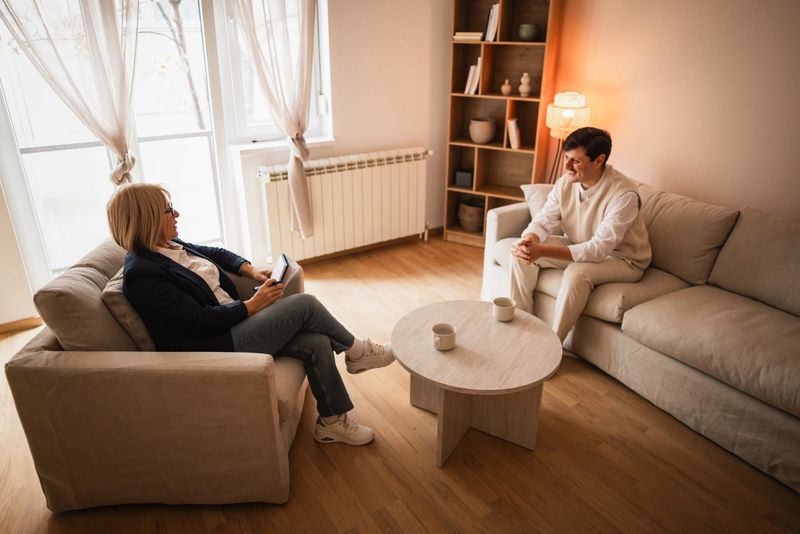 A mature female therapist and a young male client are engaged in an in-person psychotherapy session in a warm, neutral-toned office. The cozy interior features soft lighting, comfortable seating, and natural daylight streaming through sheer curtains. The scene reflects a supportive and professional mental health environment focused on emotional well-being, trust, and open communication. Ideal for concepts like counseling, mental health support, therapy, personal growth, and wellness.
