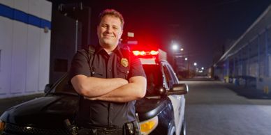 Smiling police officer stands confidently in front of a patrol car at night.