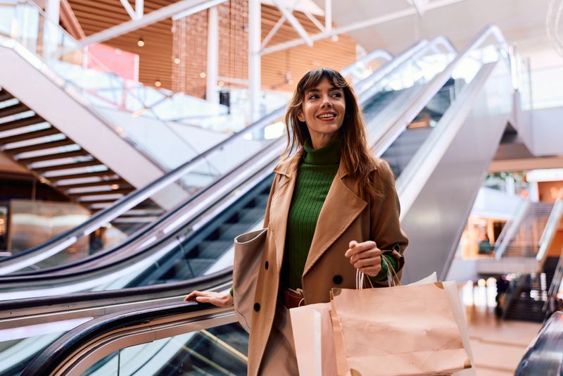 Young woman with long brown hair and shopping bags rides an escalator in a modern mall, smiling and looking away, stylish in a trench coat and green turtleneck, enjoying retail outing