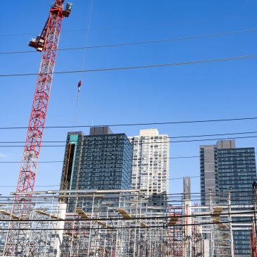 A red construction crane near modern high-rise buildings under a clear blue sky.
