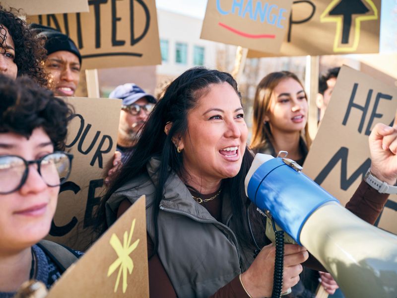 A diverse group of adults gathered outdoors holding handmade signs while a woman speaks through a megaphone during a positive community rally.