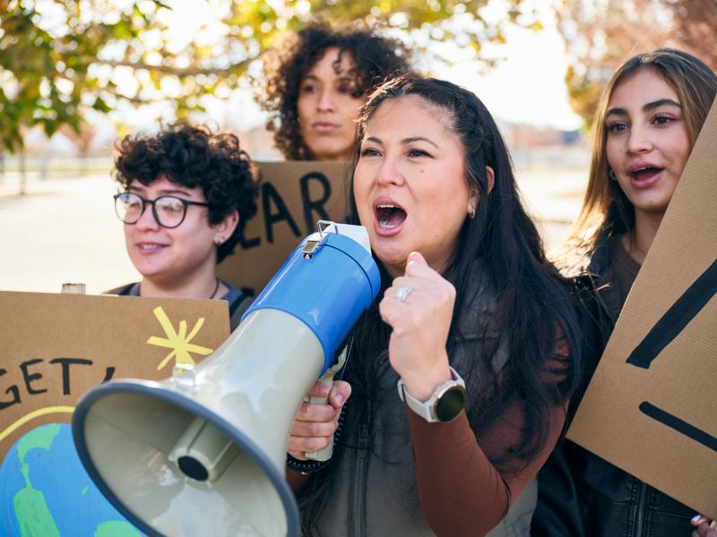 A diverse group of adults gathered outdoors holding handmade signs while a woman speaks through a megaphone during a positive community rally.