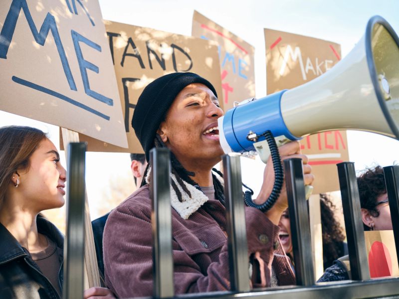 A diverse group of adults gathered outdoors holding handmade signs while a man speaks through a megaphone during a positive community rally.