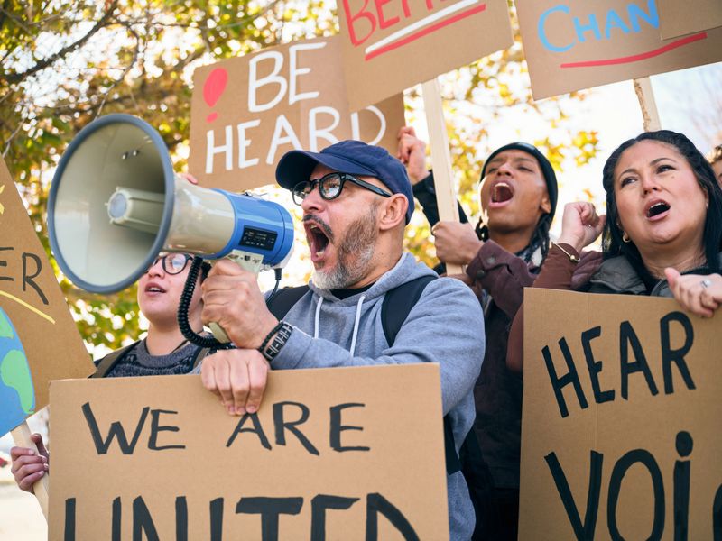 A diverse group of adults gathered outdoors holding handmade signs while a man speaks through a megaphone during a positive community rally.