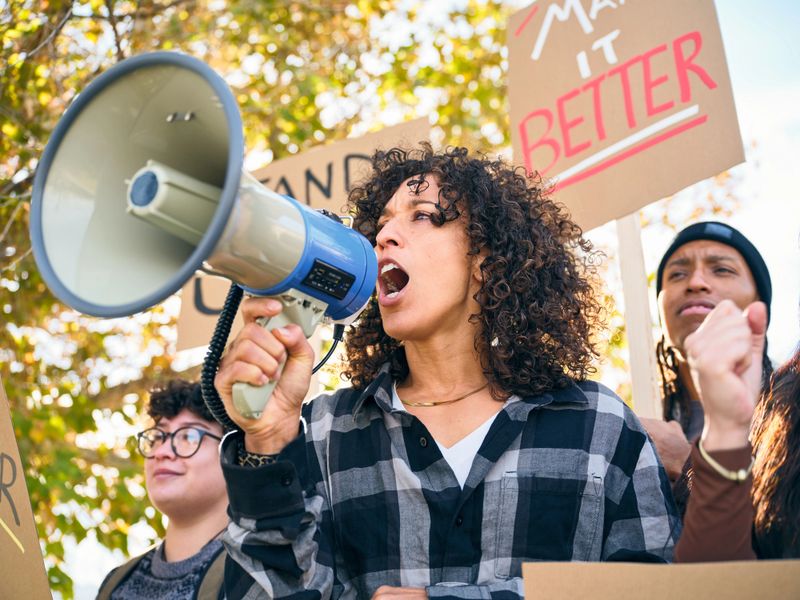 A diverse group of adults gathered outdoors holding handmade signs while a woman speaks through a megaphone during a positive community rally.