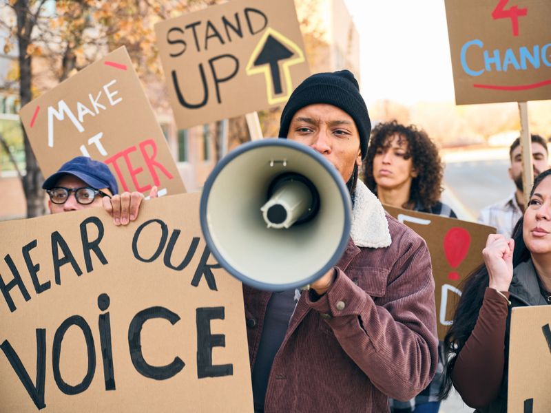 A diverse group of adults gathered outdoors holding handmade signs while a man speaks through a megaphone during a positive community rally.