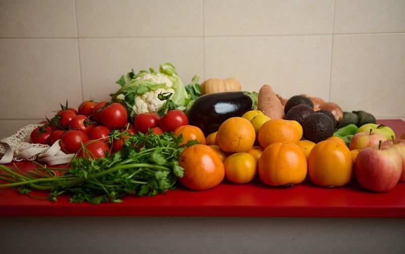 A vibrant assortment of fresh fruits and vegetables arranged on a red countertop, featuring tomatoes, oranges, apples, cauliflower, eggplant, greens, and herbs for food, market, and healthy living themes.