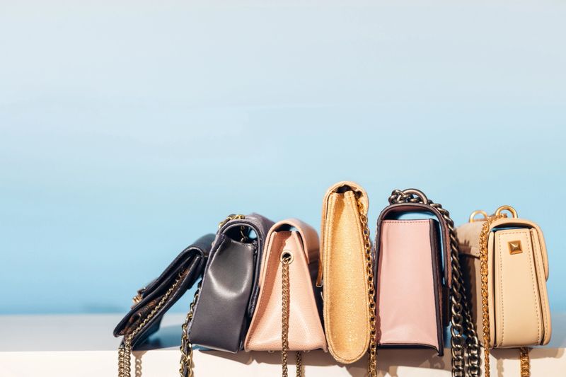 Stylish handbags arranged in a neat row on a shelf, featuring pastel tones, gold chains, and sleek designs, creating a clean and modern fashion display with organized accessory storage.
