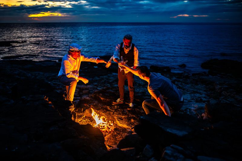 Three friends gather around a campfire on a rocky beach at dusk. The setting sun casts a warm glow, enhancing the serene atmosphere. They engage in a moment of connection, symbolizing mindfulness and mental wellbeing amidst nature's tranquility, captured with a contre-jour lighting.