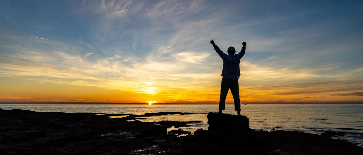 Person celebrating on rock at sunset by the sea.