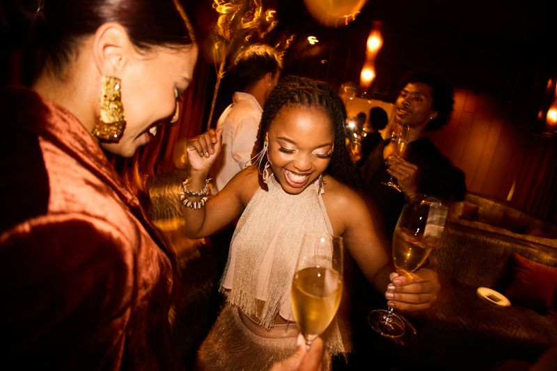 Laughing young woman in a glamorous fringed top dancing with friends in a warmly lit club environment while holding a glass of sparkling wine during a New Year’s Eve celebration.