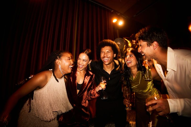 Diverse group of joyful friends laughing together during a glamorous New Year’s Eve party illuminated by soft warm lights and sparkling gold decorations.
