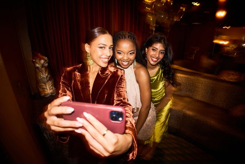 Three cheerful young women smiling warmly while taking a selfie together during a lively New Year’s Eve party featuring soft lighting and golden decorative elements.
