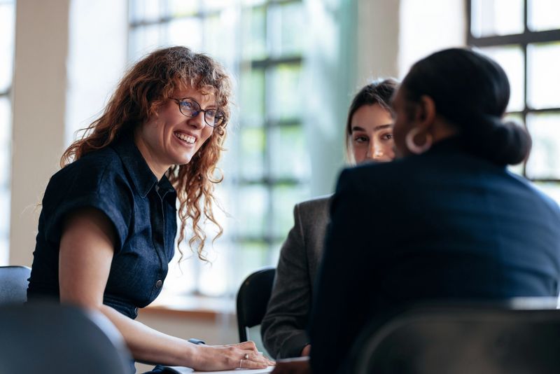 A group of smiling professionals mid-discussion around a table in a bright, modern office. The scene conveys teamwork, collaboration, and positive energy during a casual business meeting.
