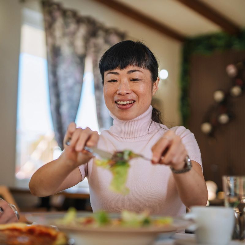Asian woman enjoying a healthy meal, eating a fresh green salad at the dining table, smiling happily and looking forward to her nutritious lunch in a bright dining room