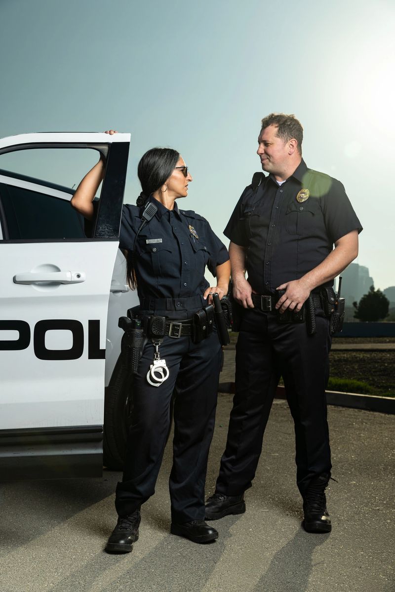 Portrait of two cops, a Southeast Asian woman and a white man, posing while standing next to a police squad car.