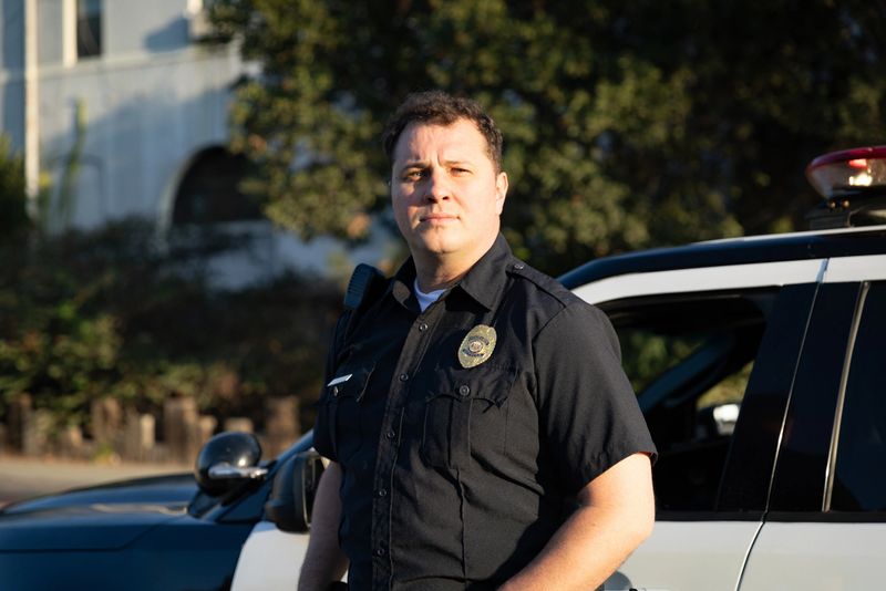 A white policeman looks off camera in front of a squad car on a sunny day.
