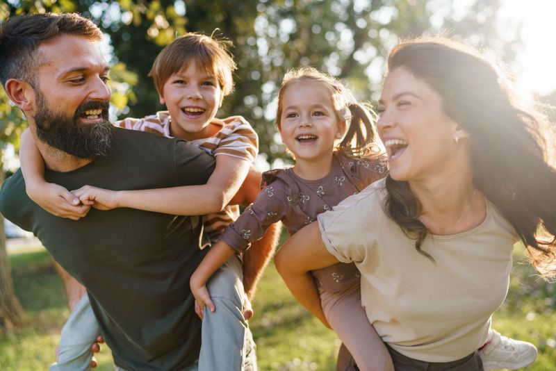 Happy family having fun together in a park, parents giving piggyback rides to their kids.