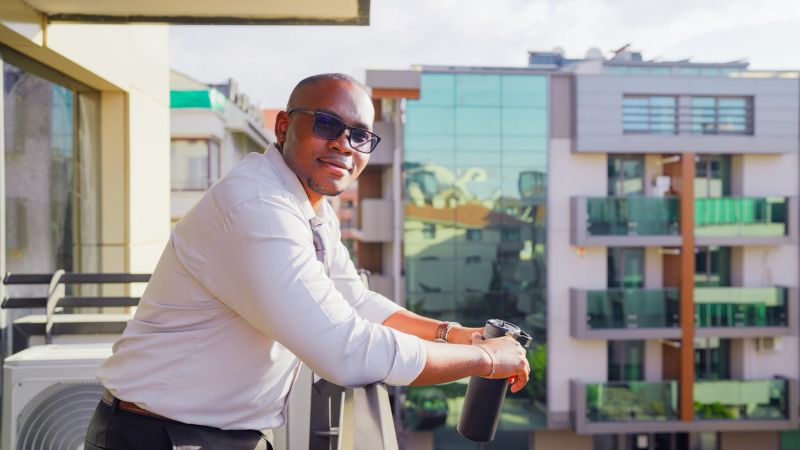 A portrait of a black, ethnic businessman taking a break from work and standing in the balcony and looking at the camera with a smile in a modern office.