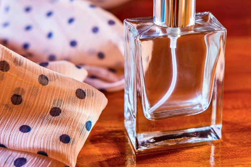 A clear glass perfume bottle with a silver cap sits beside a soft, patterned fabric, bathed in warm, inviting light.